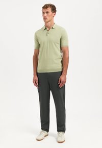 Lichtgroen polo shirt met korte mouwen en drie knopen, gecombineerd met donkergrijze broek en crèmekleurige sneakers, staand voor een witte achtergrond.