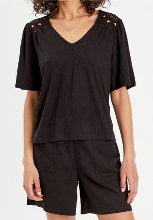 Femme portant un haut noir texturé à manches courtes avec des épaules en dentelle crochetée et un short noir, accessoirisée avec des colliers dorés superposés.