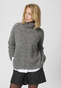 Pull en maille grise à col roulé avec une coupe ample, associé à un short en cuir noir. Le pull présente une finition texturée et un col côtelé.