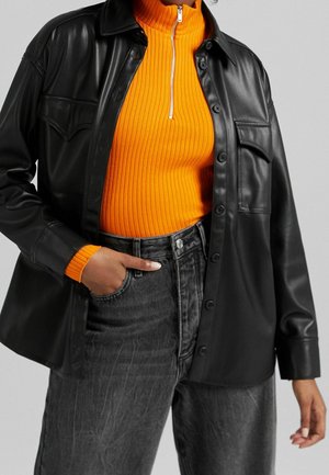 Person wearing a bright orange ribbed sweater with a zipper and a black buttoned faux leather jacket over faded black high-waisted jeans.