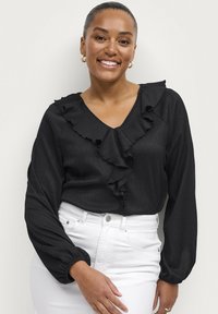 Blusa nera con scollatura a V e dettagli arricciati sul davanti. Maniche lunghe con polsini arricciati. Indossata con jeans bianchi a vita alta.