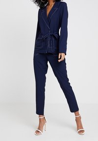 Žena v námořnicky modrém sáčku s páskem, sladěných kalhotách a bílých vysokých podpatcích, stojící na jednobarevném pozadí.