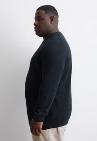 Dunkel gestrickter Pullover in einem einfarbigen Marineblau. Verfügt über einen Rundhalsausschnitt, lange Ärmel sowie gerippte Bündchen und Saum. Getragen mit beigen Hosen.