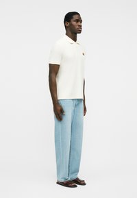 Hombre de pie mirando hacia la derecha, vistiendo un polo blanco de manga corta, jeans de pierna ancha azul claro y sandalias marrón oscuro sobre un fondo blanco liso.