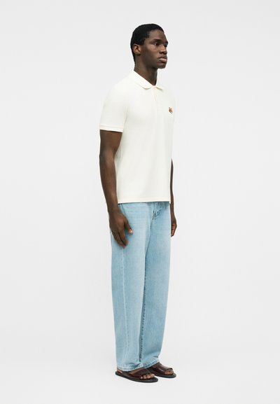 Un homme est debout, tourné vers la droite, portant un polo blanc à manches courtes, un jean bleu clair à jambes larges, et des sandales marron foncé sur un fond blanc uni.