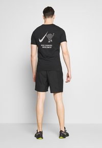 Schwarzes Nike-T-Shirt mit einem grafischen Druck auf dem Rücken, kombiniert mit schwarzen Shorts und schwarzen Laufschuhen mit gelben Akzenten.