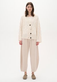 Cardigan a maglia cable cream con bottoni neri sopra un top beige chiaro e pantaloni a gamba larga. Texture morbida con una vestibilità rilassata e colori neutri.