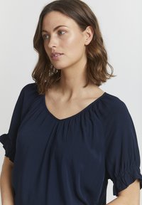 Top blu navy realizzato in tessuto morbido, con scollatura a V, cuciture delle spalle arricciate e maniche corte voluminose.