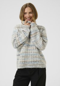 Kobieta uśmiechająca się i trzymająca kołnierz przytulnego, wielokolorowego swetra dzianinowego, ubrana w czarne spodnie, na tle jasnego, jednolitego tła.