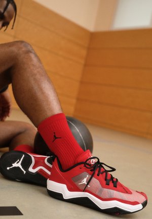 Červené sportovní boty s černými akcenty; syntetický svršek s prodyšnými panely. Nosené s odpovídajícími červenými ponožkami se znakem. V pozadí basketbal.
