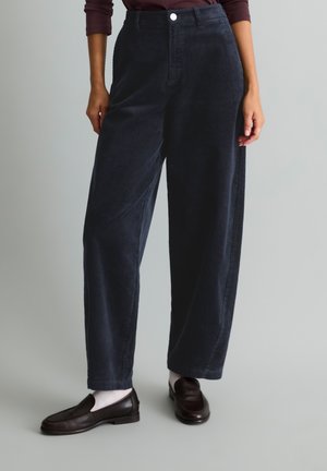 Persoon die donkerblauwe wijde ribfluwelen broek draagt met een knoopsluiting in de taille, witte sokken en donkerbruine loafers, staand tegen een effen achtergrond.