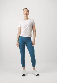 Sportos öltözet világos rózsaszín, testhez álló pólóval és türkiz leggings-szel, fehér sportcipővel kombinálva. Simább anyag, testreszabott szabás.