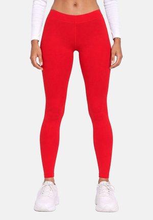 Femme portant des leggings rouges vifs taille haute, un crop top blanc à manches longues et des baskets blanches, debout devant un fond uni.