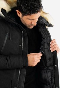 Parka noire avec capuche en fourrure, poches zippées et poignets côtelés ; elle présente une fermeture éclair brillante et un intérieur matelassé.