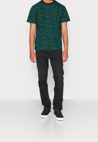 T-shirt verde a fantasia geometrica, abbinata a jeans neri e sneaker neri con suola bianca, in piedi su uno sfondo neutro.