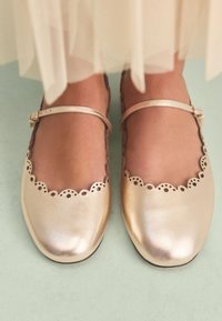 Ballerines Mary Jane en cuir doré avec un bord festonné et des détails découpés le long du rebord. Texture douce, bout rond et bride de cheville ajustable.