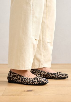 Płaskie buty w zwierzęcy wzór w panterkę z czarną podeszwą, wykonane z tkaniny, z ozdobną kokardką z przodu, zestawione z beżowymi spodniami o szerokich nogawkach.