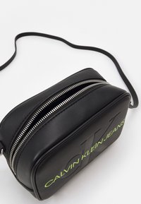 Czarna skórzana torba crossbody o gładkiej fakturze, z zamknięciem na zamek błyskawiczny i wyraźnym logo "CALVIN KLEIN JEANS" w zielonym kolorze po boku.