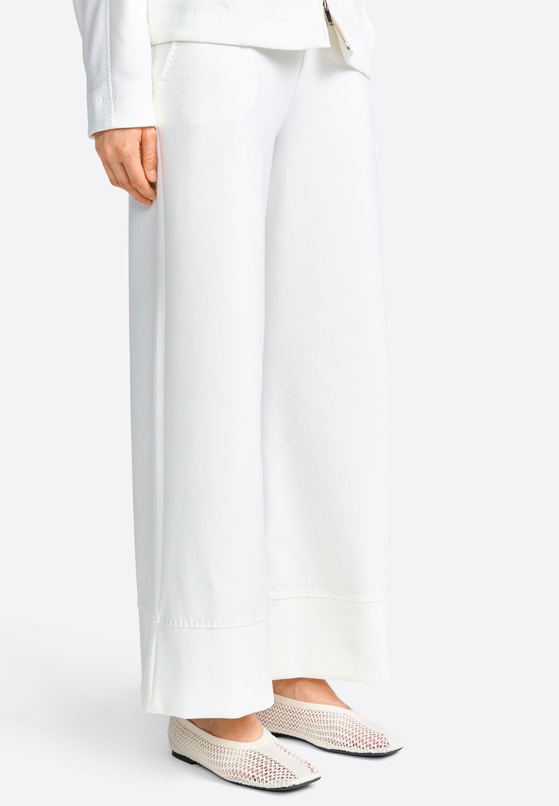 Persona con pantalones blancos de pierna ancha y zapatos blancos de malla sin cordones, de pie sobre un fondo claro.