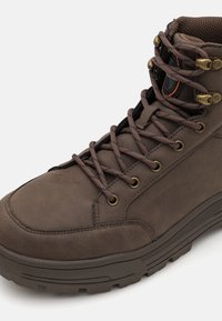 Bota de senderismo marrón con cordones, hecha de un material sintético texturizado. Presenta ojales de metal y una suela resistente para mayor tracción.