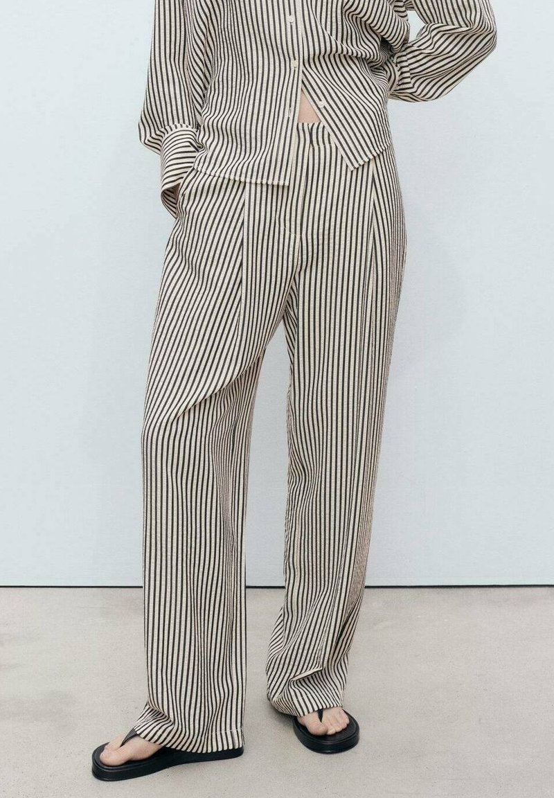 Personne portant un pantalon beige ample et un haut assorti avec des rayures verticales noires, debout avec une main sur la hanche et portant des tongs noires.
