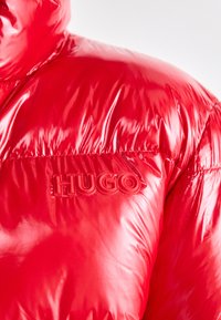 Red puffer jacket with a glossy finish, featuring a textured logo patch that reads "HUGO" on the left side of the chest.