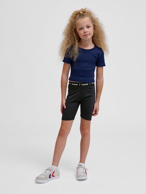 Ung pige med krøllet hår iført en mørkeblå t-shirt, sorte shorts med "hummel" talje, og hvide sneakers, stående mod en ensfarvet baggrund.