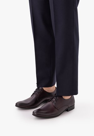 Sötétbarna bőrből készült, fűzős elegáns cipők, szabott tengerészkék nadrággal, fehér háttér előtt.