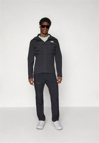 Μαύρο τζάκετ με φερμουάρ και επενδυμένη μπροστινή όψη, σετ με στενά μαύρα παντελόνια. Το μοντέλο φοράει ανοιχτοπράσινο πουκάμισο και γυαλιά ηλίου, με γκρι παπούτσια.