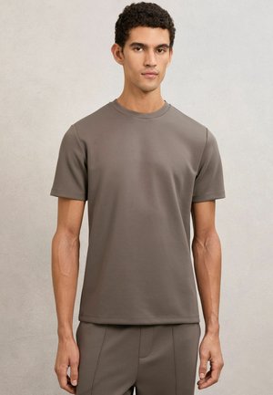 Ung mand med kort krøllet hår iført en ensfarvet taupe t-shirt med korte ærmer og matchende bukser foran en neutral baggrund.