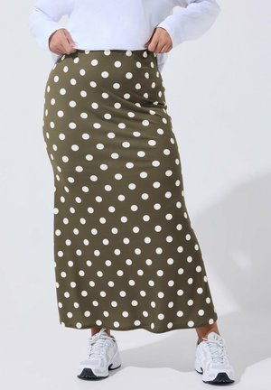 Persoon draagt een olijfgroene rok met witte polka dots en witte sneakers, terwijl hij een wit overhemd dat in de rok is gestopt, rechtzet.