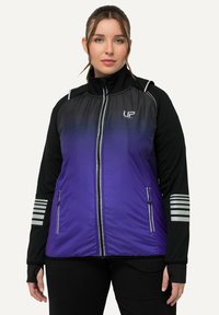 Kvindelig iført en lilla og sort sportjakke med lynlås, der har reflekterende striber på ærmerne og UP Sports-logo på brystet, står med ansigtet vendt fremad.