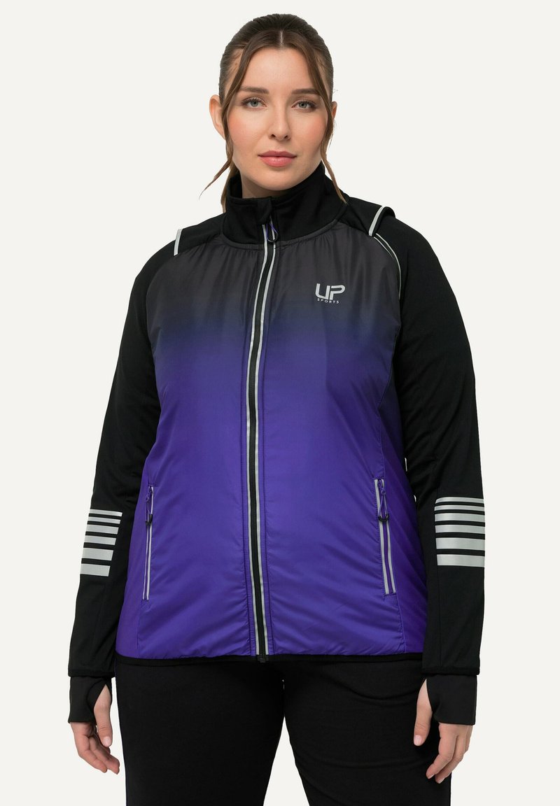 Donna che indossa una giacca sportiva zip-up nera e viola con strisce riflettenti sulle maniche e il logo UP Sports sul petto, in piedi di fronte.