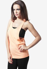 Tank top v broskvové barvě s širokými ramínky a volným střihem, nošený přes černé přiléhavé tričko, v kombinaci s černými legínami. Hladká textura látky.