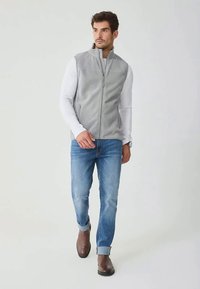 Graue Fleeceweste mit Frontreißverschluss, getragen über einem weißen Langarmshirt, kombiniert mit blauen Jeans und braunen Stiefeletten.