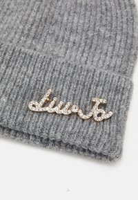 Beanie in maglia a costine grigia con una spilla metallica dorata che spellera "live" adornata con strass trasparenti, caratterizzato da una texture morbida e uno stile aderente.