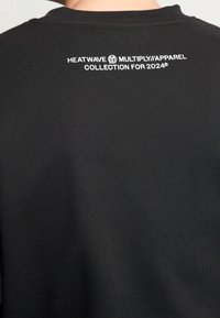 Camiseta de algodón negra con cuello redondo y una etiqueta impresa en blanco en la parte posterior que dice "COLECCIÓN DE ROPA HEATWAVE MULTIPLY PARA 2024."
