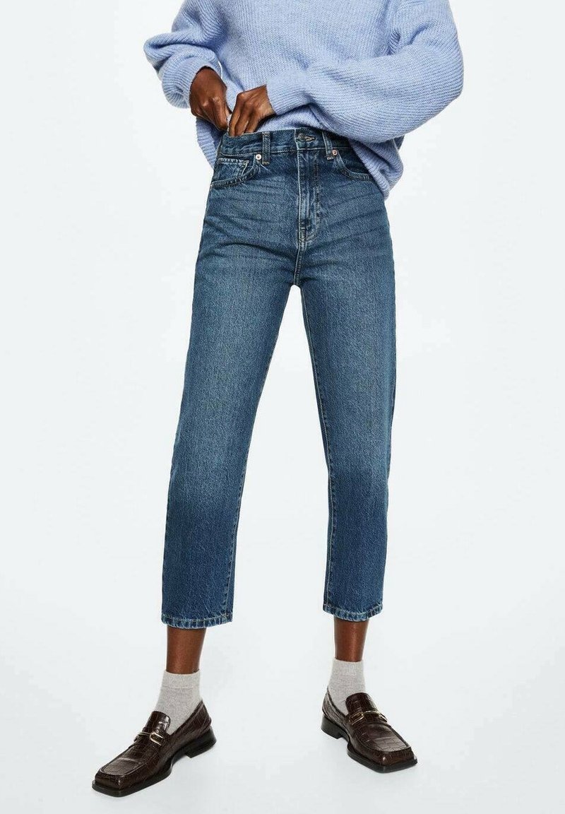 Personne portant un jean court bleu, des chaussettes gris clair, des mocassins marron foncé et un pull en tricot bleu clair ajustant la taille du jean.