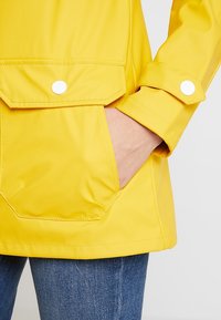 Chubasquero amarillo con un acabado brillante, botones a presión en los puños y en la tapa del bolsillo, y un bolsillo delantero. Combinado con jeans de mezclilla azules.