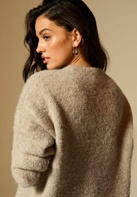 Beżowy puszysty sweter o luźnym fasonie, opadających ramionach i prążkowanych mankietach. Modelka prezentuje bok, z miękką teksturą włosów i biżuterią.