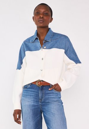 Donna con capelli corti che indossa un cardigan blu e bianco con bottoni, jeans in denim blu e una cintura marrone con fibbia decorativa.