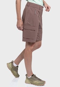 Braune Cargo-Shorts mit lockerem Schnitt, ausgestattet mit seitlichen Taschen und einem Reißverschlussdetail. Kombiniert mit olivgrünen Sportschuhen.