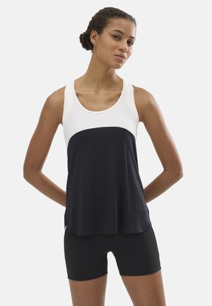 Mujer con el cabello recogido en un moño, vistiendo una camiseta sin mangas bicolor (blanca y negra) y pantalones cortos negros, de pie con las manos detrás de la espalda.