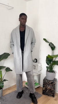 Gray wool coat with open front and large lapels, worn over a black shirt. Paired with gray jeans and black shoes. Indoor setting with plants.