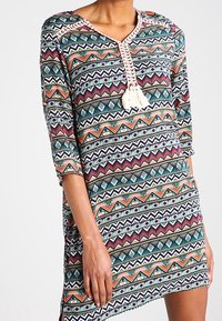 Mönstrad klänning i flerfärgad tyg med geometriska mönster. V-ringning med virkad knytning och tofsar. Trekvartsärmar och avslappnad passform.