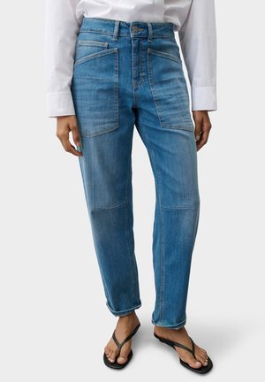 Persoon die lichtblauwe spijkerbroek draagt met grote voorzakken, opgerolde boorden, een wit shirt met lange mouwen en zwarte slippers.