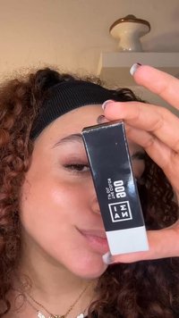 Svart rektangulär förpackning med vit botten, med en logotyp och text. Klar plastkork, hålls nära ansiktet med lockigt hår synligt.