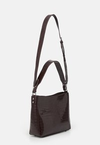 Tummanruskea krokotiilinnahkakuvioitu nahkalaukku, jossa on leveä runko, lyhyt olkahihna ja hillityt hopeanväriset metallikoristeet.