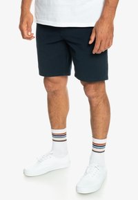 Shorts bleu marine en coton avec une coupe classique, portés avec des baskets blanches et des chaussettes mi-mollet rayées aux accents rouges, bleus et gris.