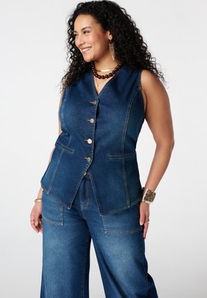 Lachende vrouw met krullend haar, gekleed in een mouwloze donkerdenimvest met gouden knopen en bijpassende jeans met wijde pijpen, opgedirkt met sieraden.
